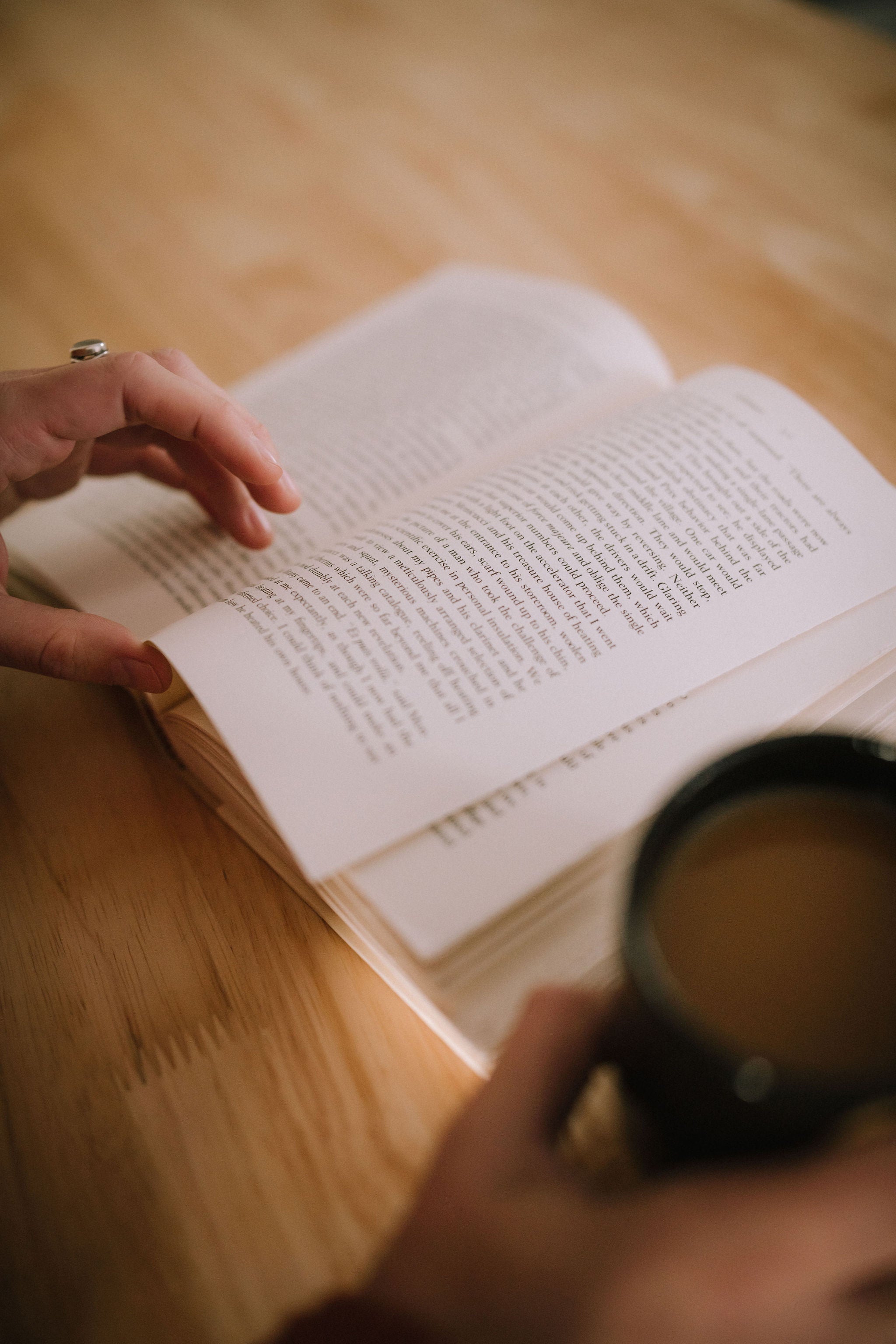 One hand turns the page of an open book, with a cup of tea in the other hand.